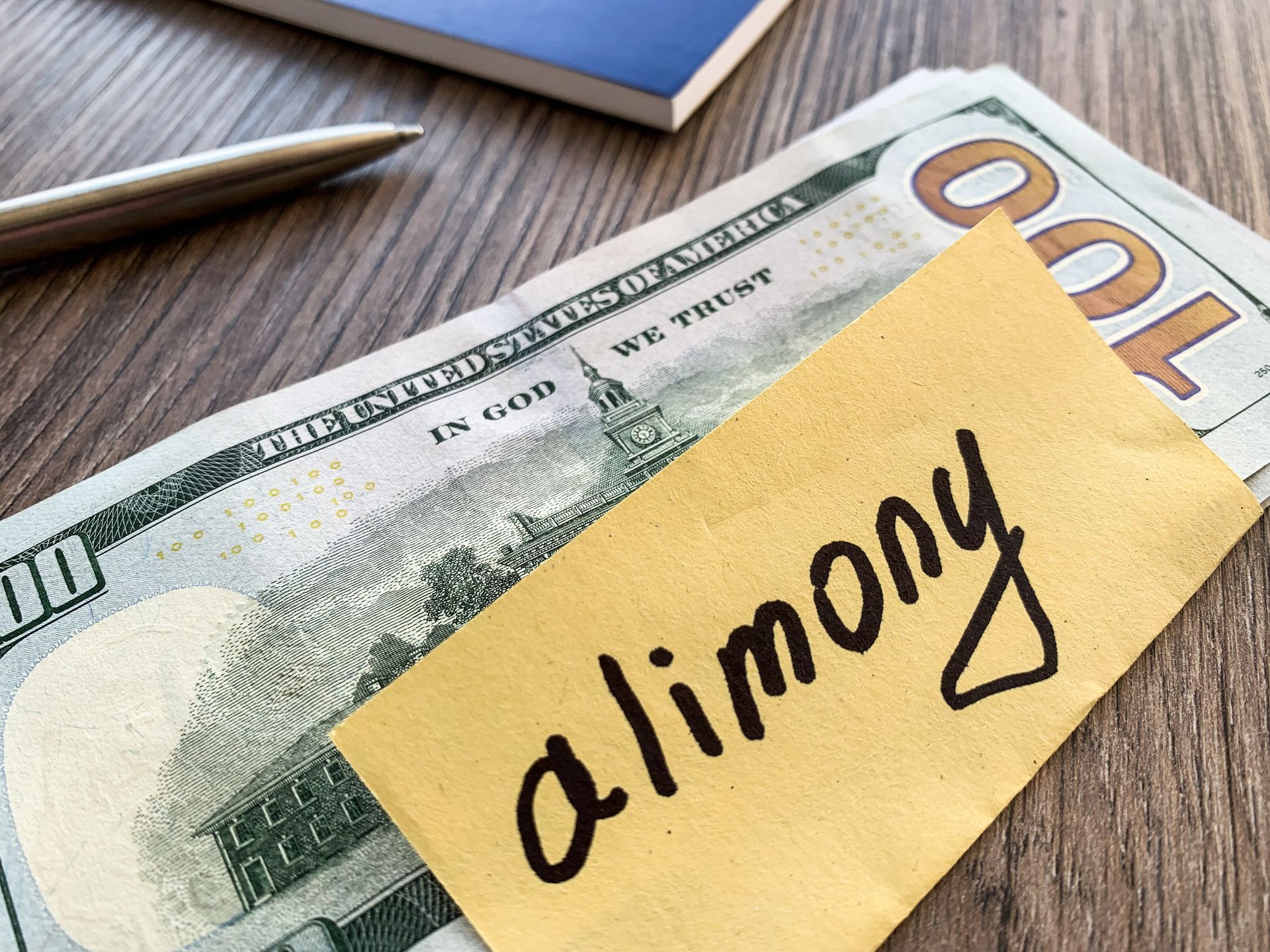 Sticky note labeled alimony resting on hundred dollar bills beside a pen on a wooden desk.