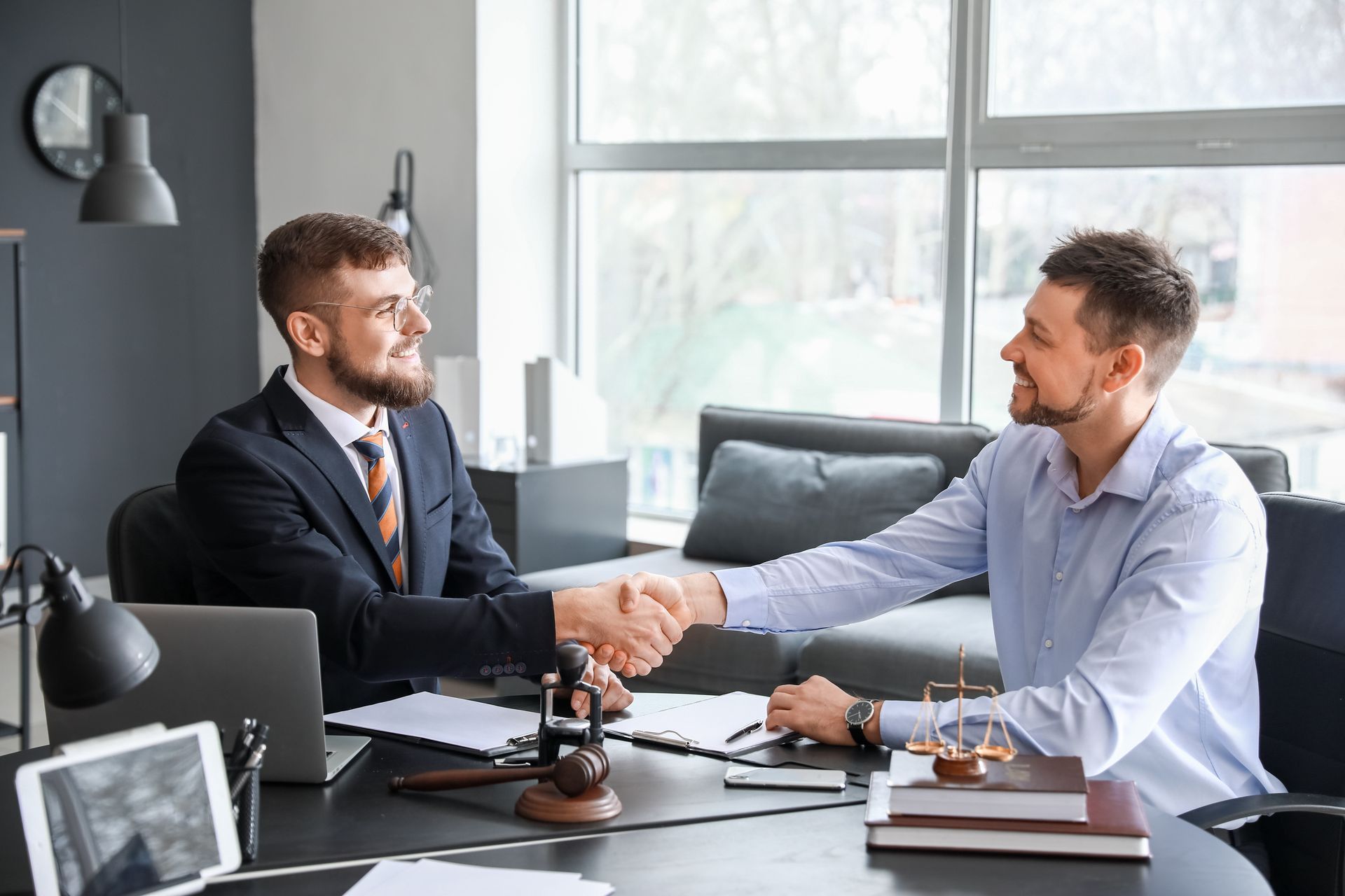 Two professionals shaking hands across desk with gavel and books in a legal office setting.