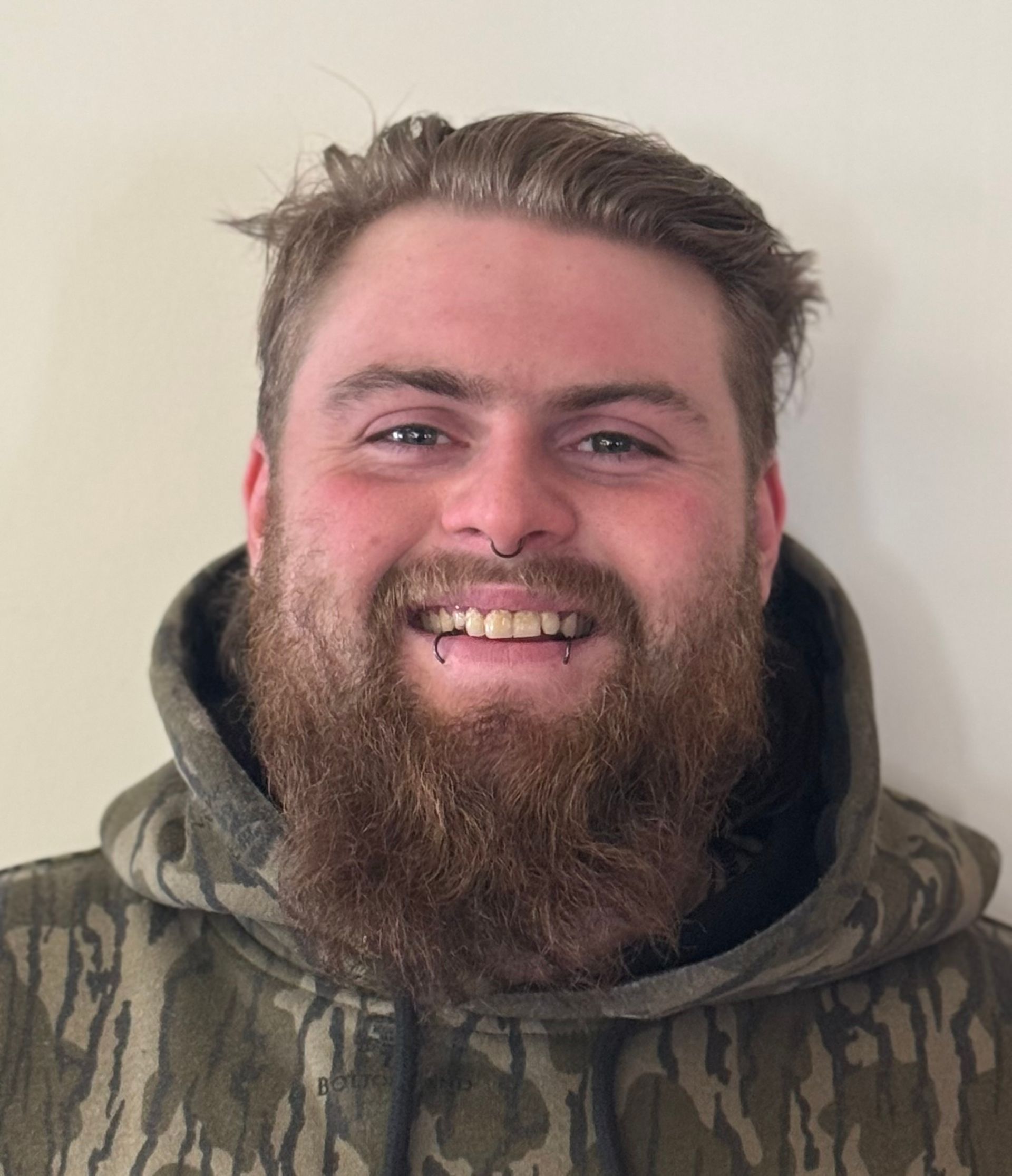 A man with a beard is wearing a camouflage hoodie and smiling.