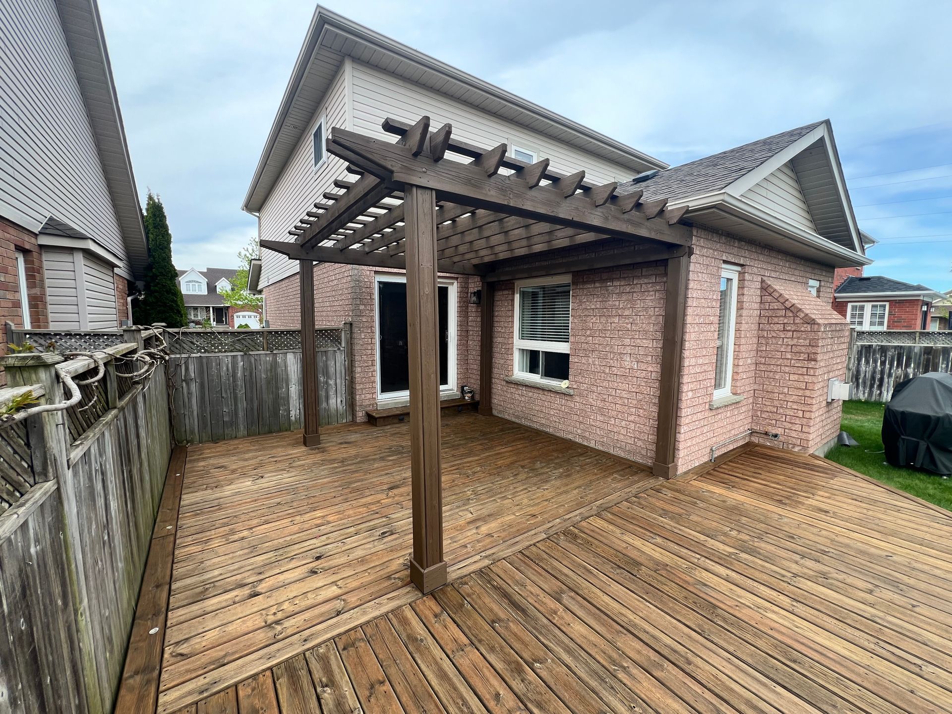 Wooden deck with pergola attached to a brick house. Brown wood, overcast sky.