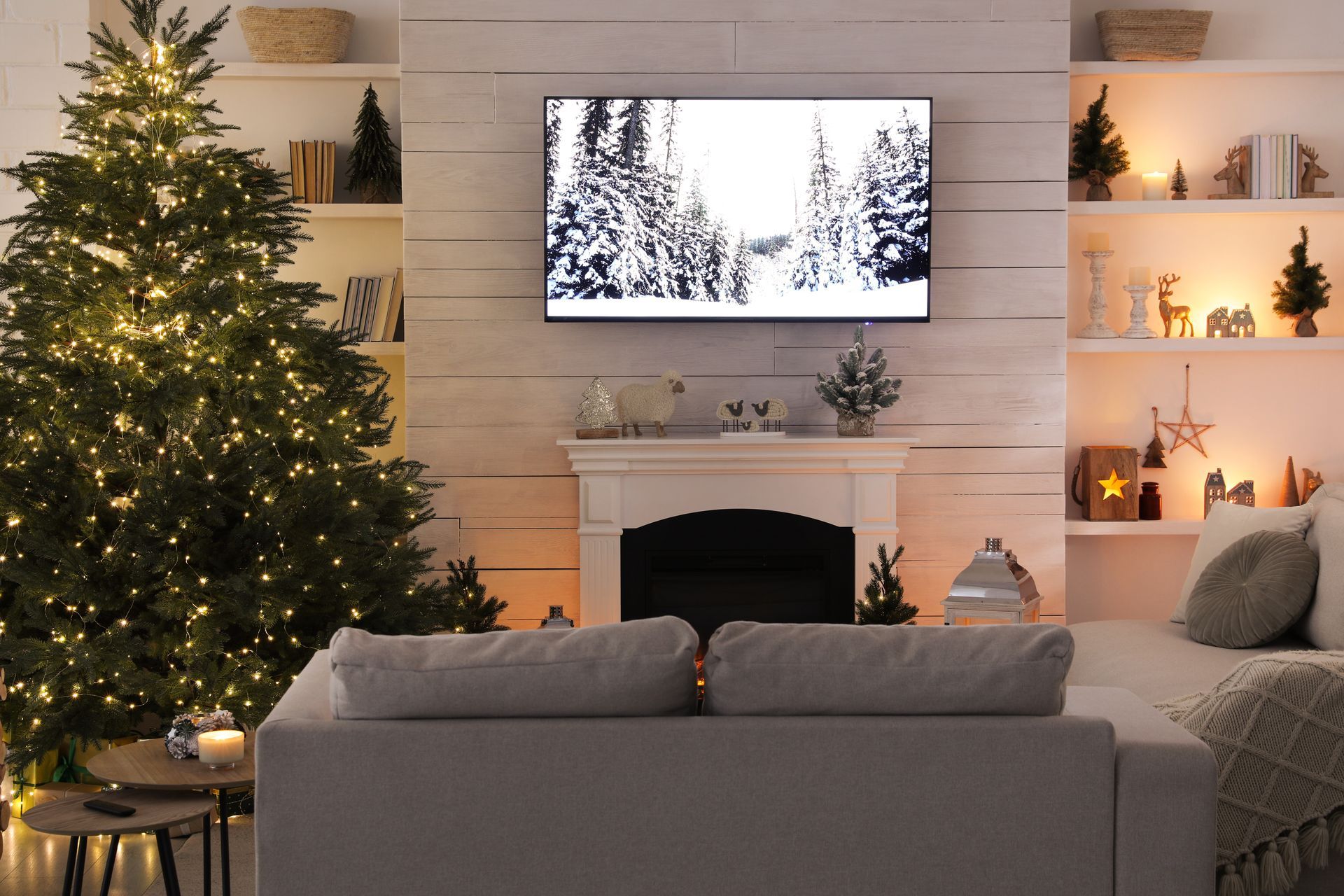 Living room decorated for Christmas with a lit tree, white sofa, and TV showing a snowy forest.