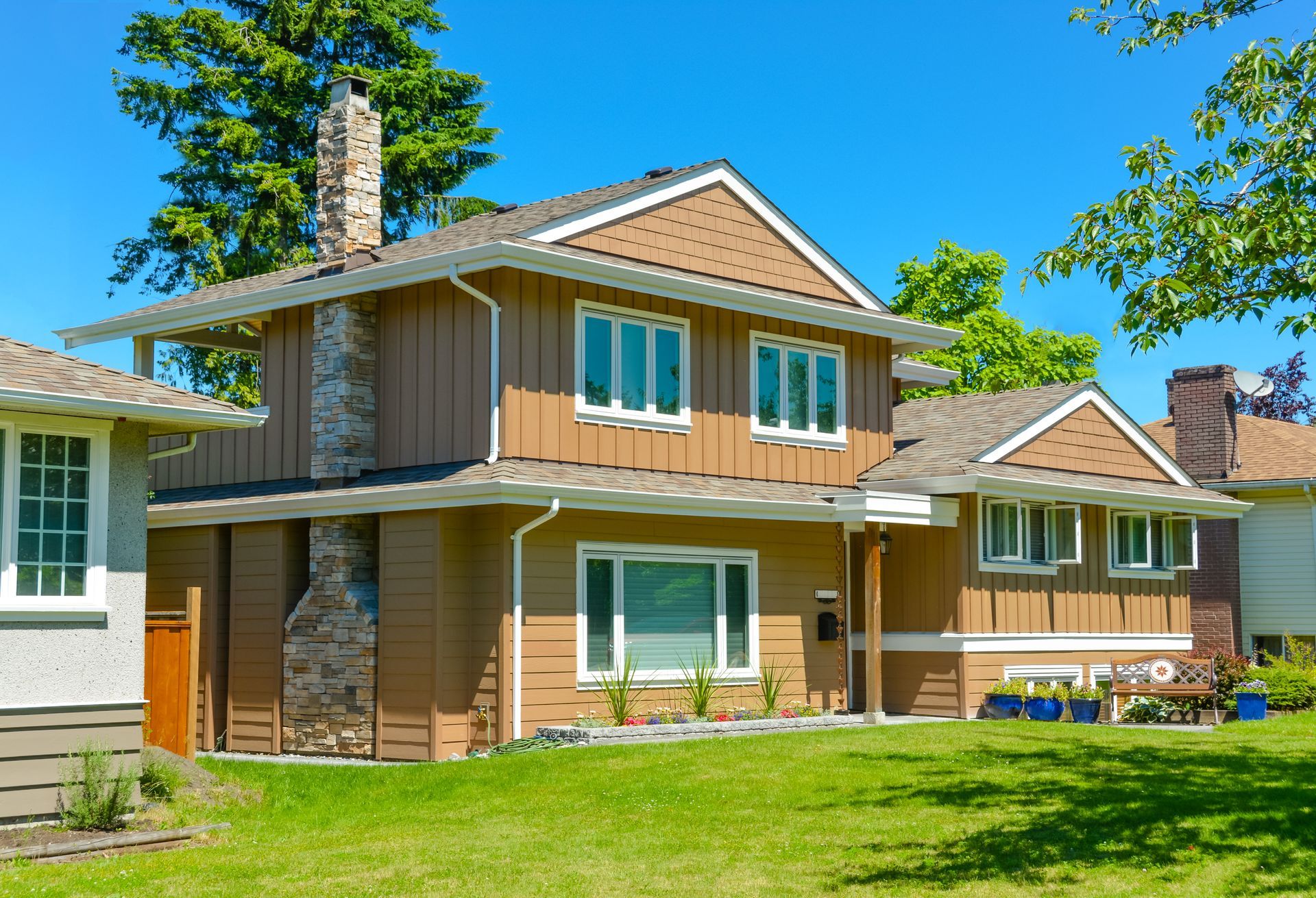 Two-story brown house with white trim, a stone chimney, and green lawn under a blue sky.