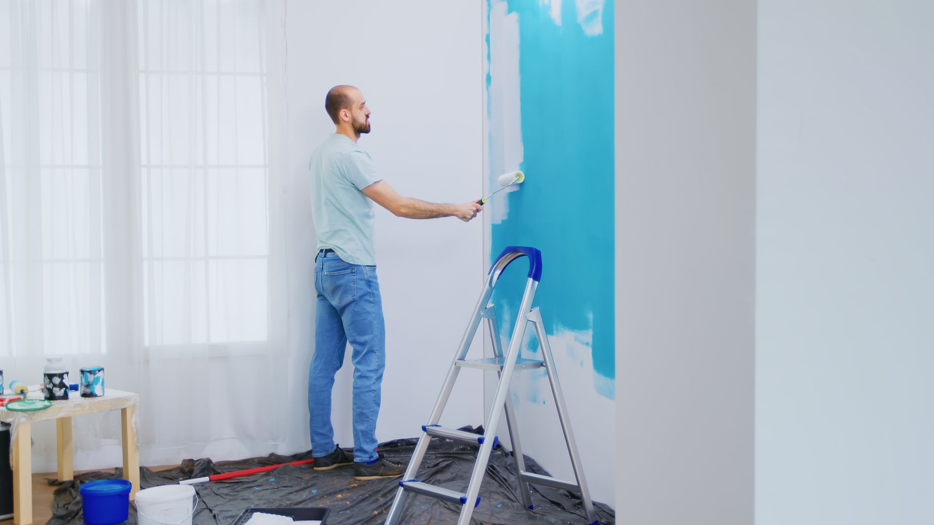 A man is standing on a ladder painting a wall blue.