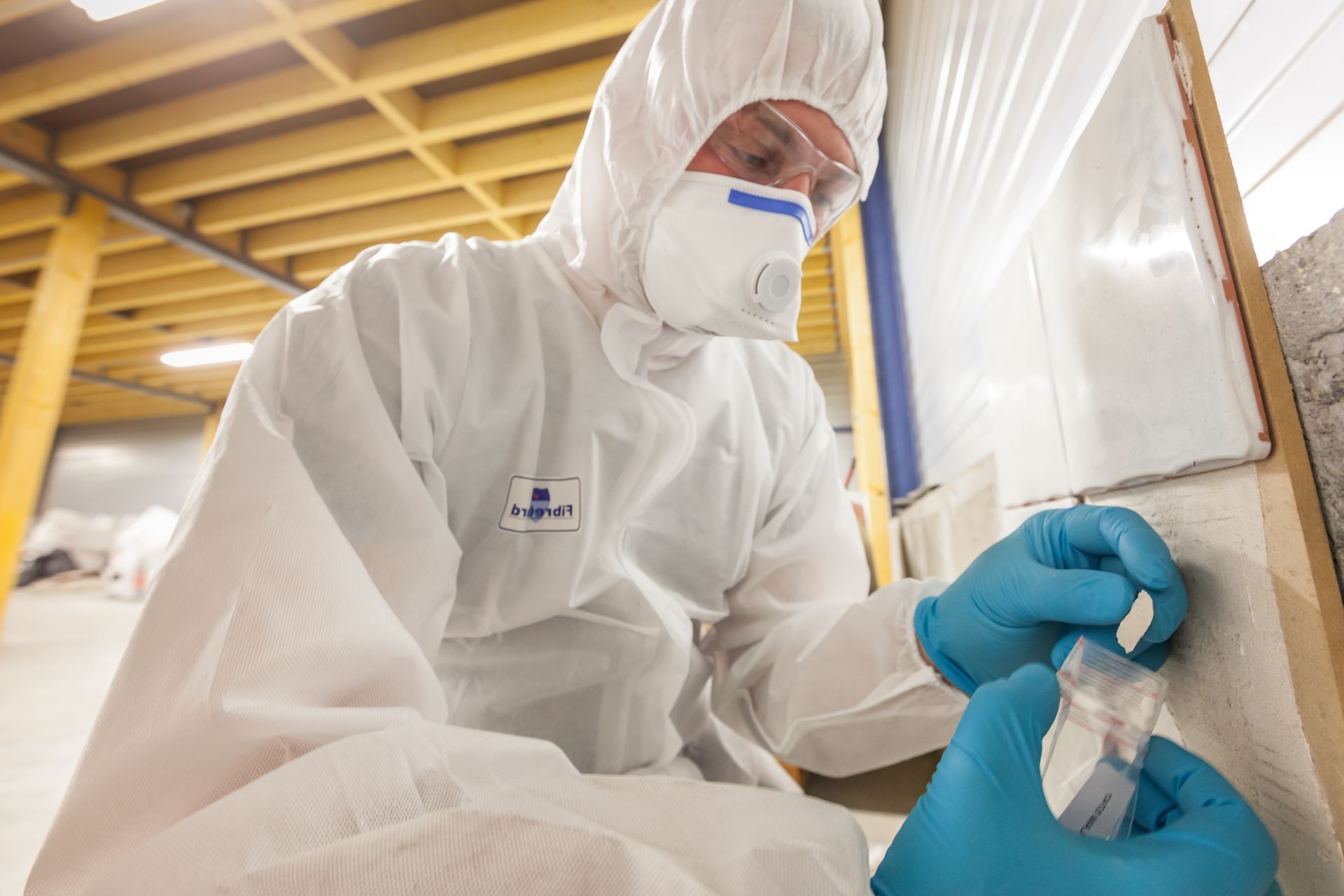 Person in a white hazmat suit and blue gloves taking a sample in a basement.