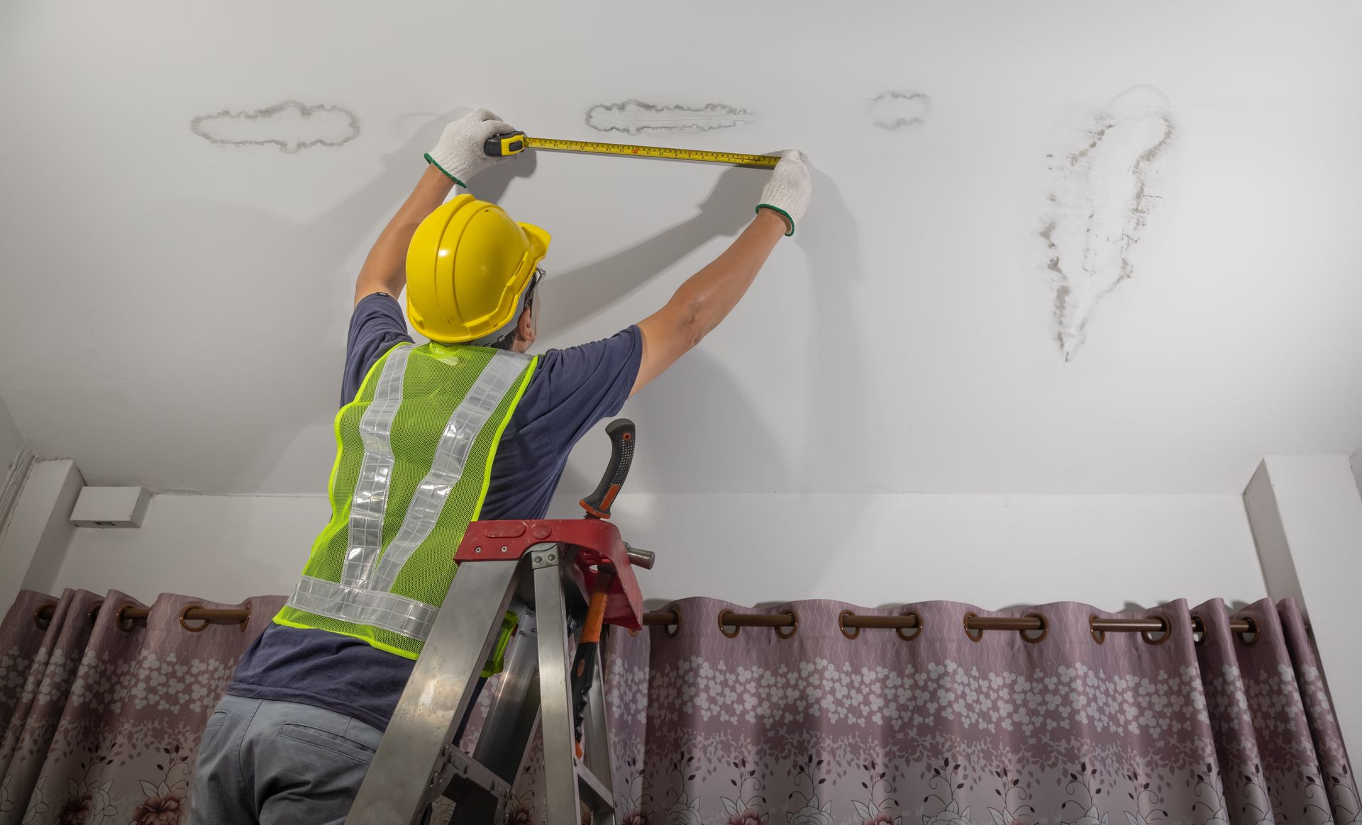 Person in a safety vest and hard hat on a ladder measures water damage on a ceiling, near patterned curtains.