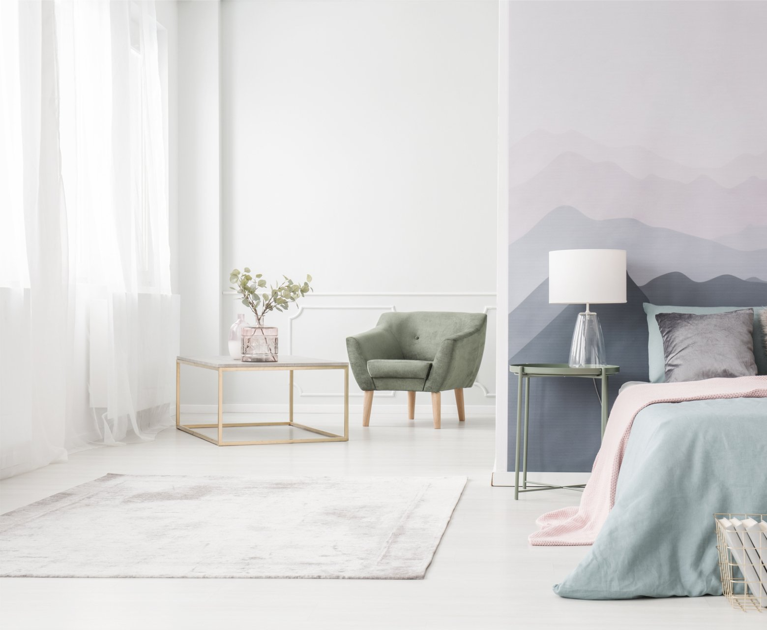 Pastel coloured bedroom showing bed, sidetable and lamp, with soft chair and table in front of opaque curtains.