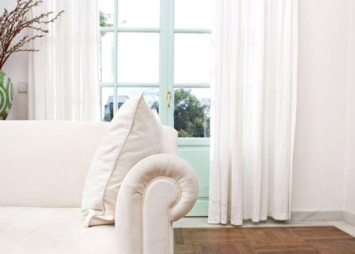 White formal couch in forefront of loungeroom. Pale gree french doors framed by open soft white curtains.