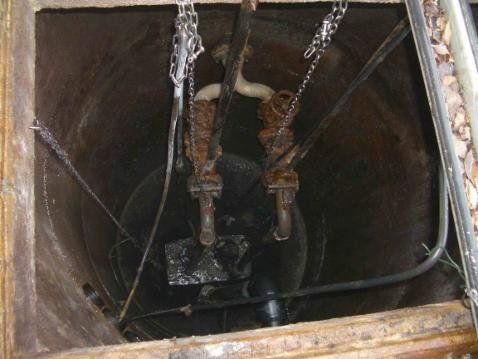 Sumpfpumpe in einer dunklen, kreisrunden Grube. Ketten und Rohre sind sichtbar.