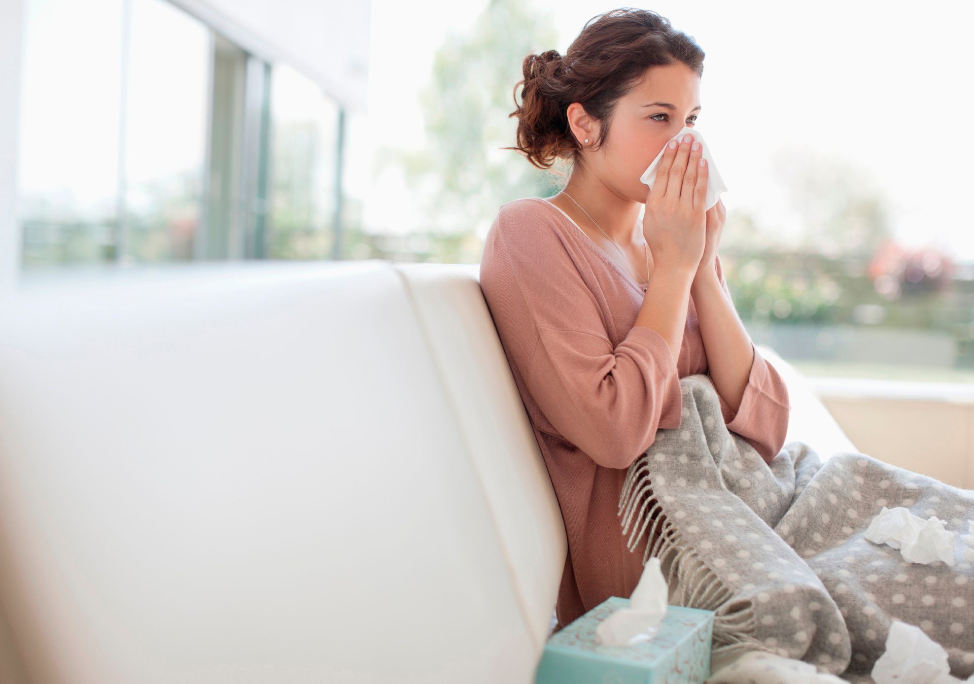 A woman is sitting on a couch blowing her nose into a napkin.