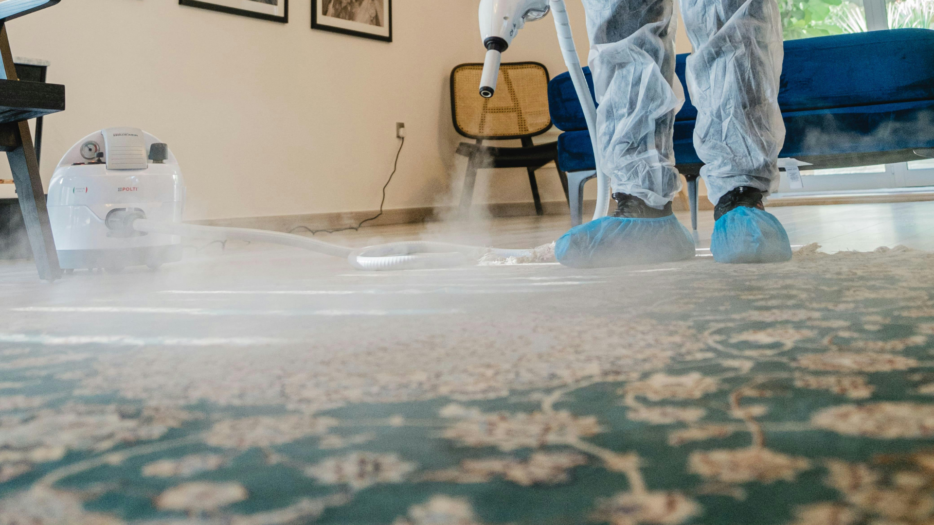 A person in a protective suit is cleaning a carpet with a steam cleaner.