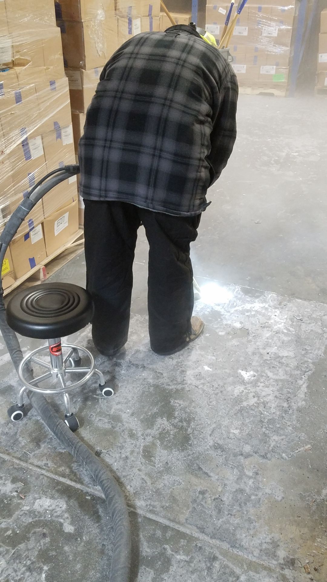Person in plaid shirt bends over a concrete floor covered in white powder; a chair and hose are nearby.