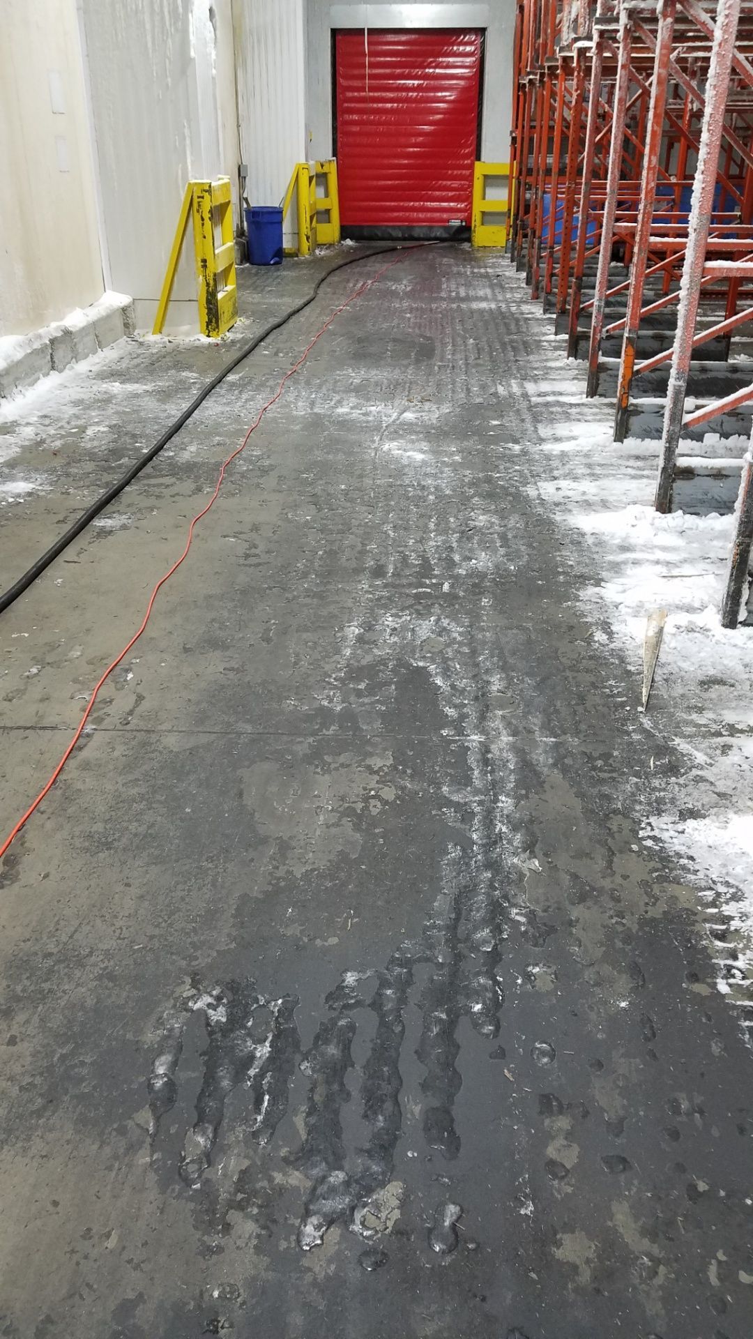 A long, wet, and icy concrete industrial corridor leading to a red shutter door. Yellow safety cones line the sides.