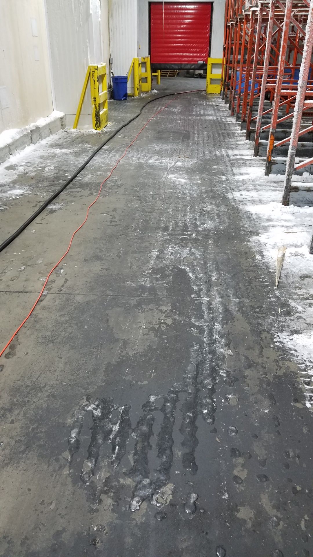 Warehouse aisle with ice and snow. Red door in background. Ladders, yellow poles, and black hose present.