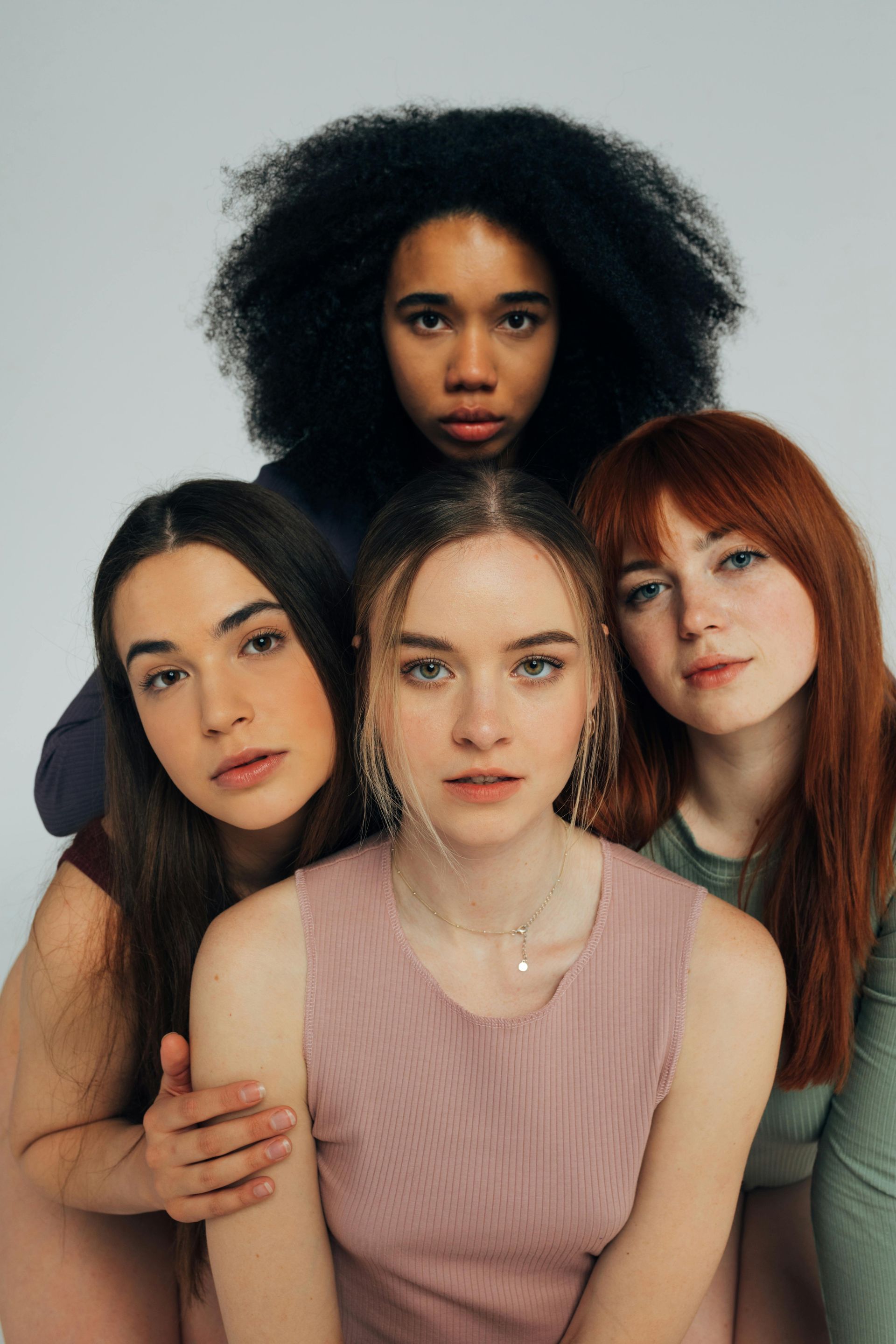 Four diverse young women posing together. One with dark curly hair, others with straight hair, wearing pastel tops, looking at the camera.