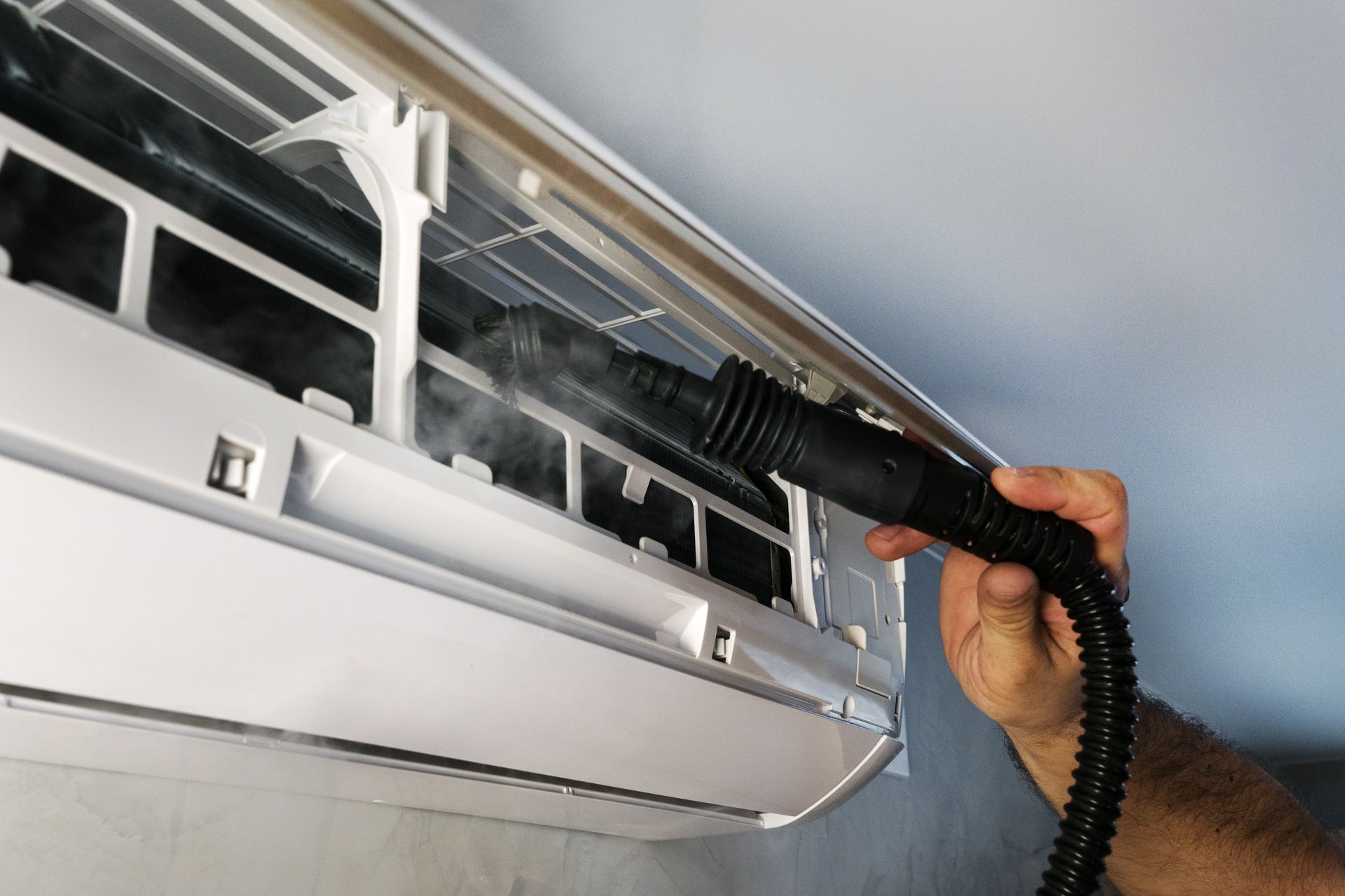 Une personne nettoie un climatiseur mural à l'aide d'une buse, créant de la vapeur visible.