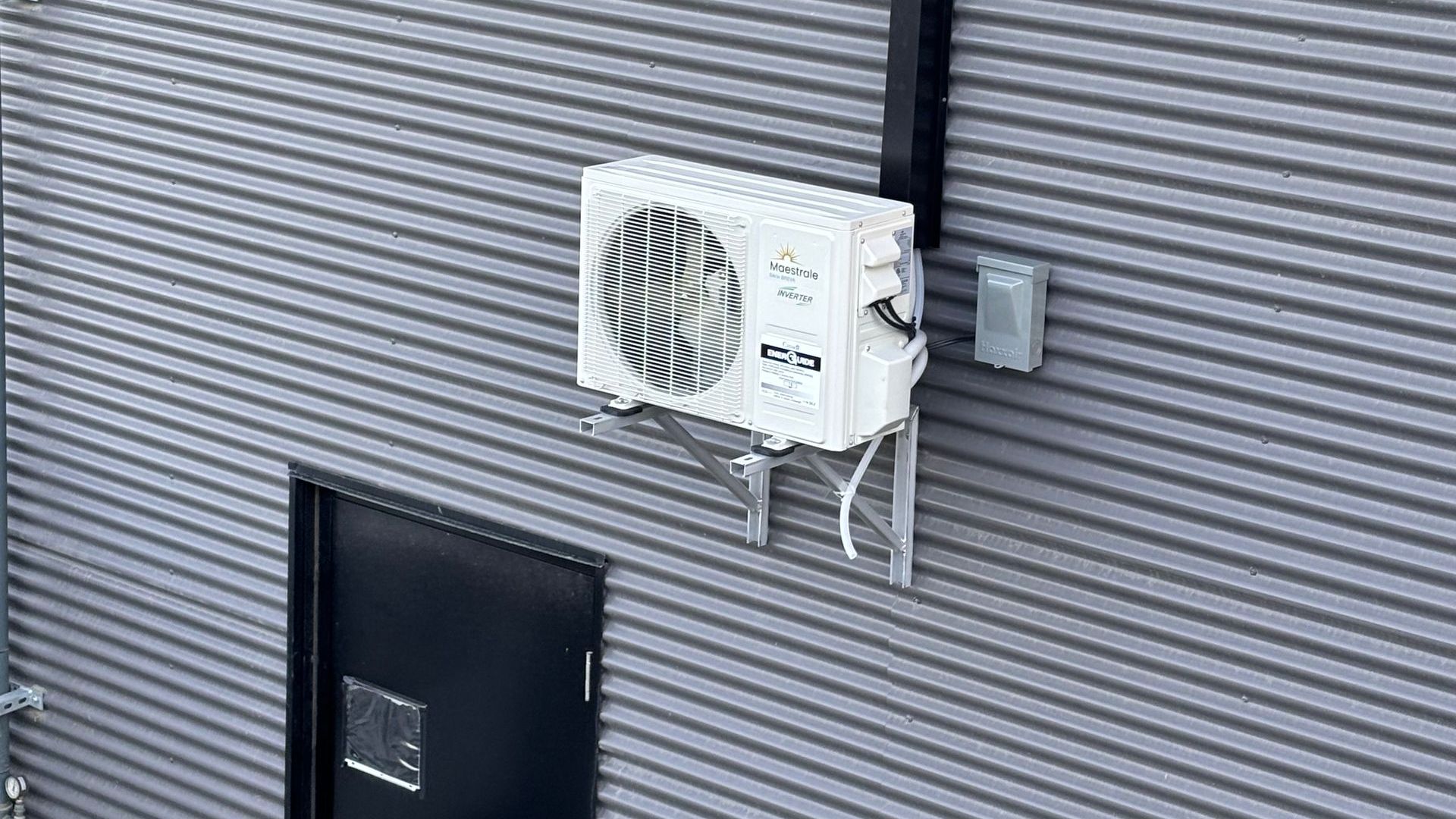 Unité de climatisation blanche fixée sur un mur en tôle ondulée grise, à côté d'une porte noire.
