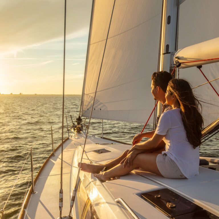 A couple sits on the deck of a sailboat, looking out at the ocean during a golden sunset.