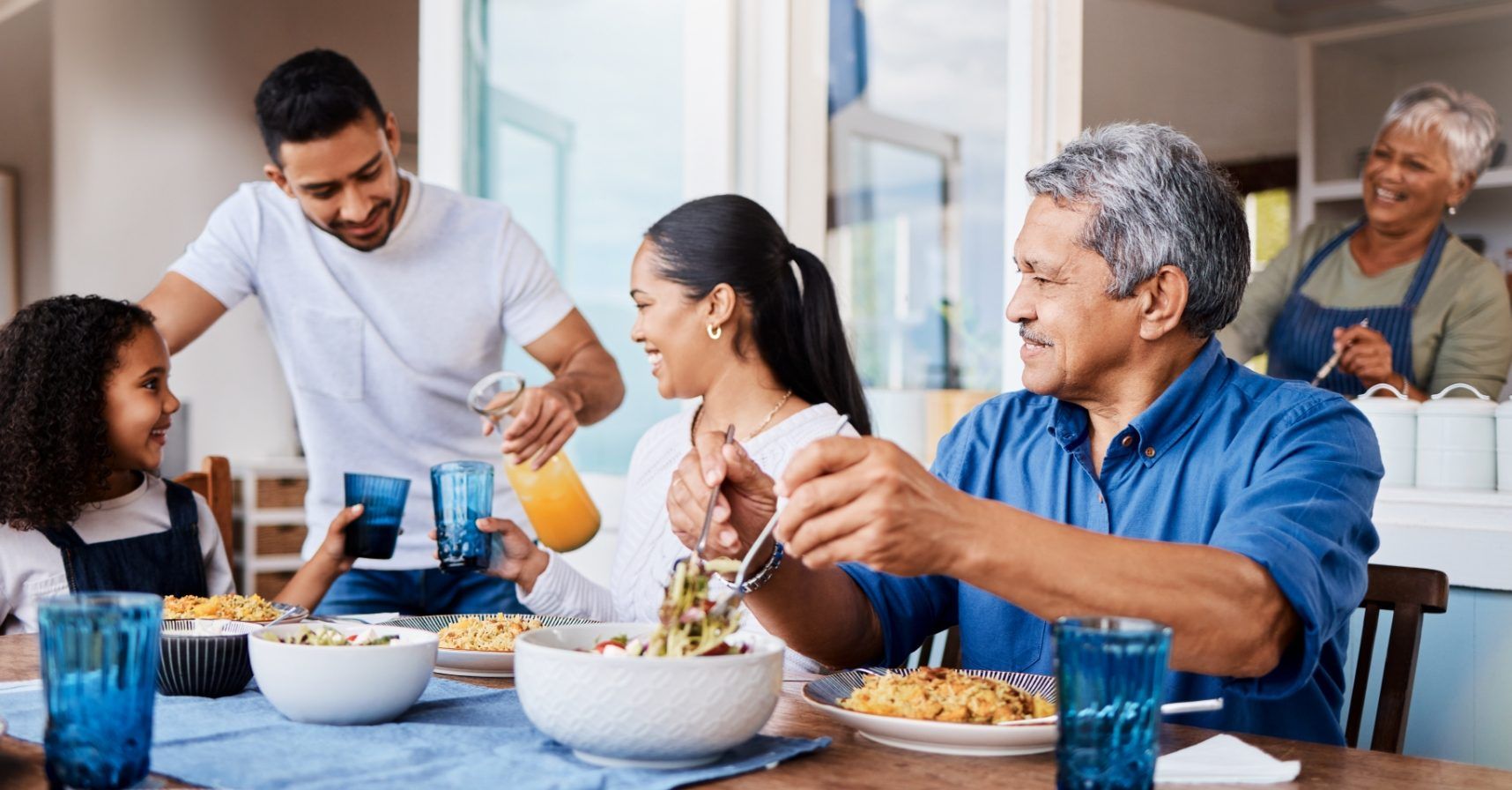 A multi-generational family eats lunch together at a dining table, laughing and pouring orange juice.