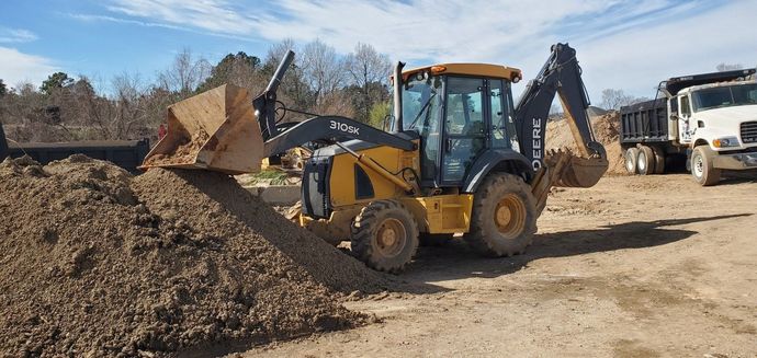 A yellow backhoe dumping dirt onto a pile, with a dump truck in the background, under a blue sky. John Deere 310SK backhoe