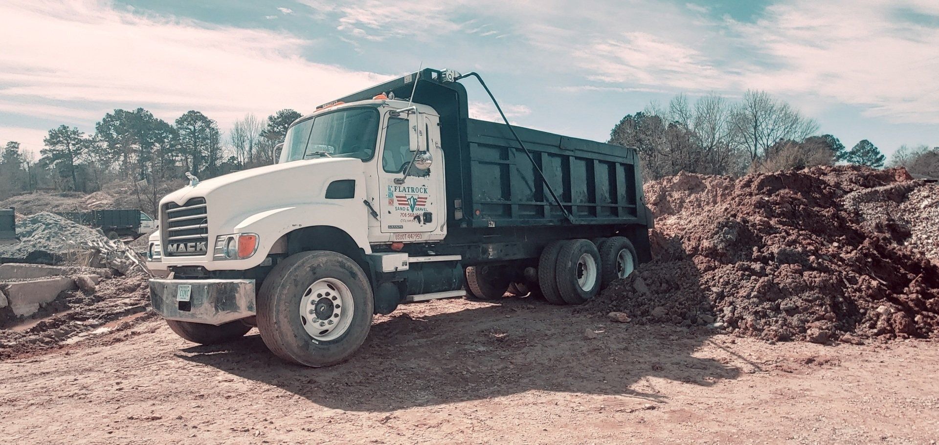 A white Mack dump truck sits on dirt, ready to be loaded, with piles of material nearby. Flatrock dump truck