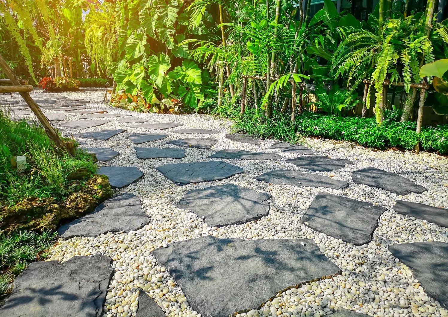 Stone pathway through lush garden, gray flagstones set in white gravel. Green foliage surrounds. Stone path in lush garden
