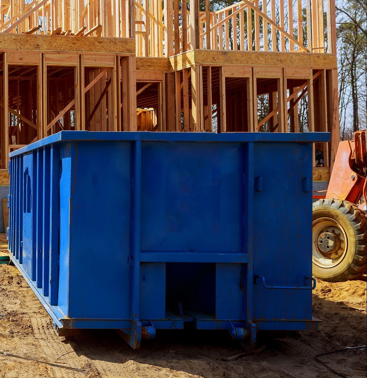 Blue construction dumpster in front of a partially built wooden house.