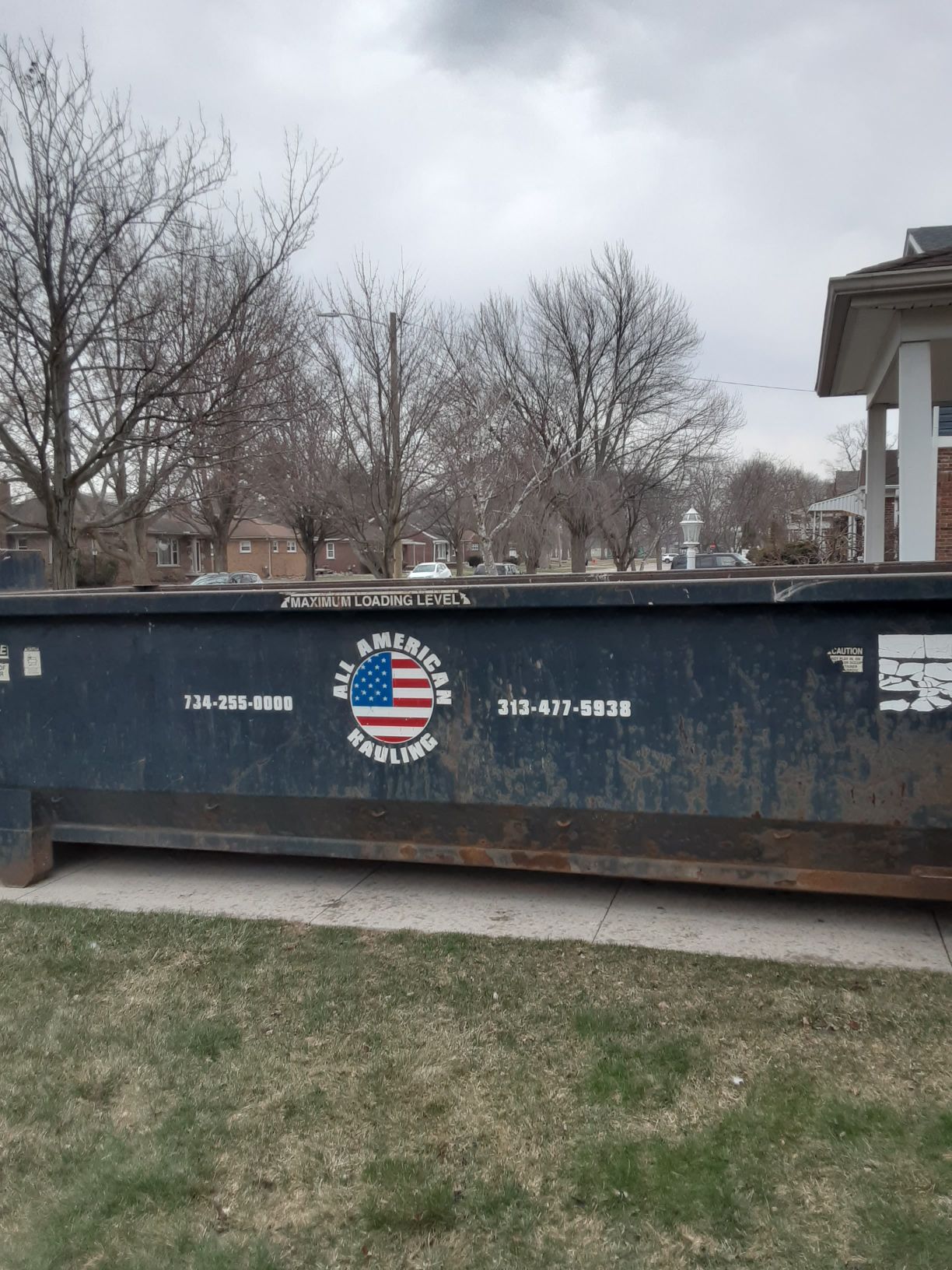 A dark blue roll-off dumpster sits on a grassy lawn near a parked truck and trailer on a sunny day.