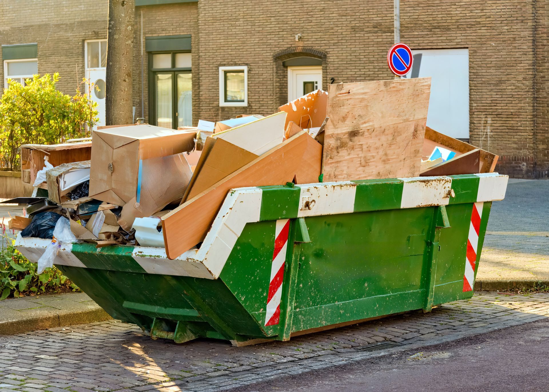 Green dumpster filled with discarded wood, cardboard, and trash, near a brick building.
