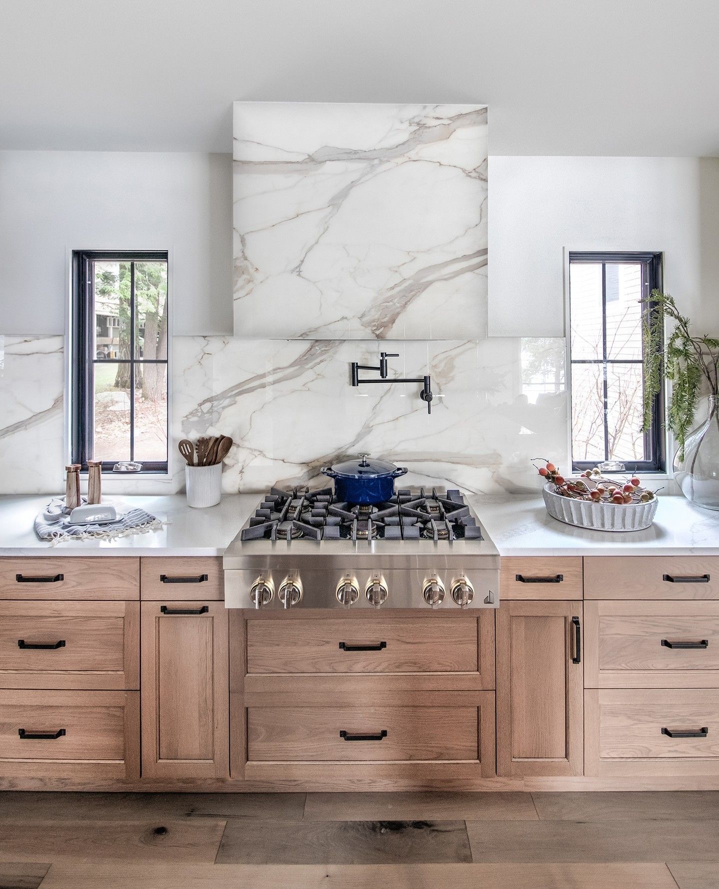 Modern kitchen with wood cabinetry, marble backsplash, central gas range, and black-framed windows on both sides