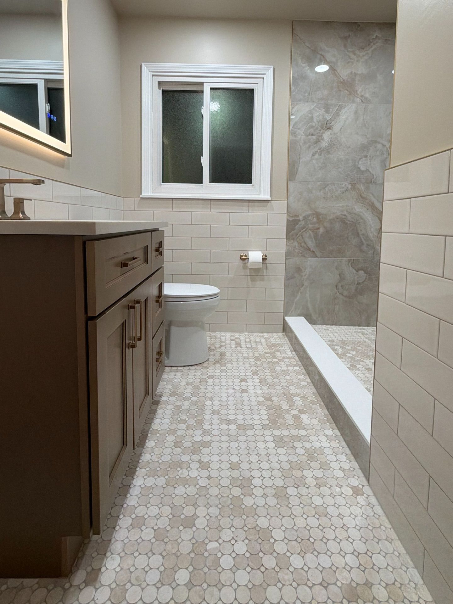 Narrow beige bathroom with vanity, toilet, shower ledge, and a small window.
