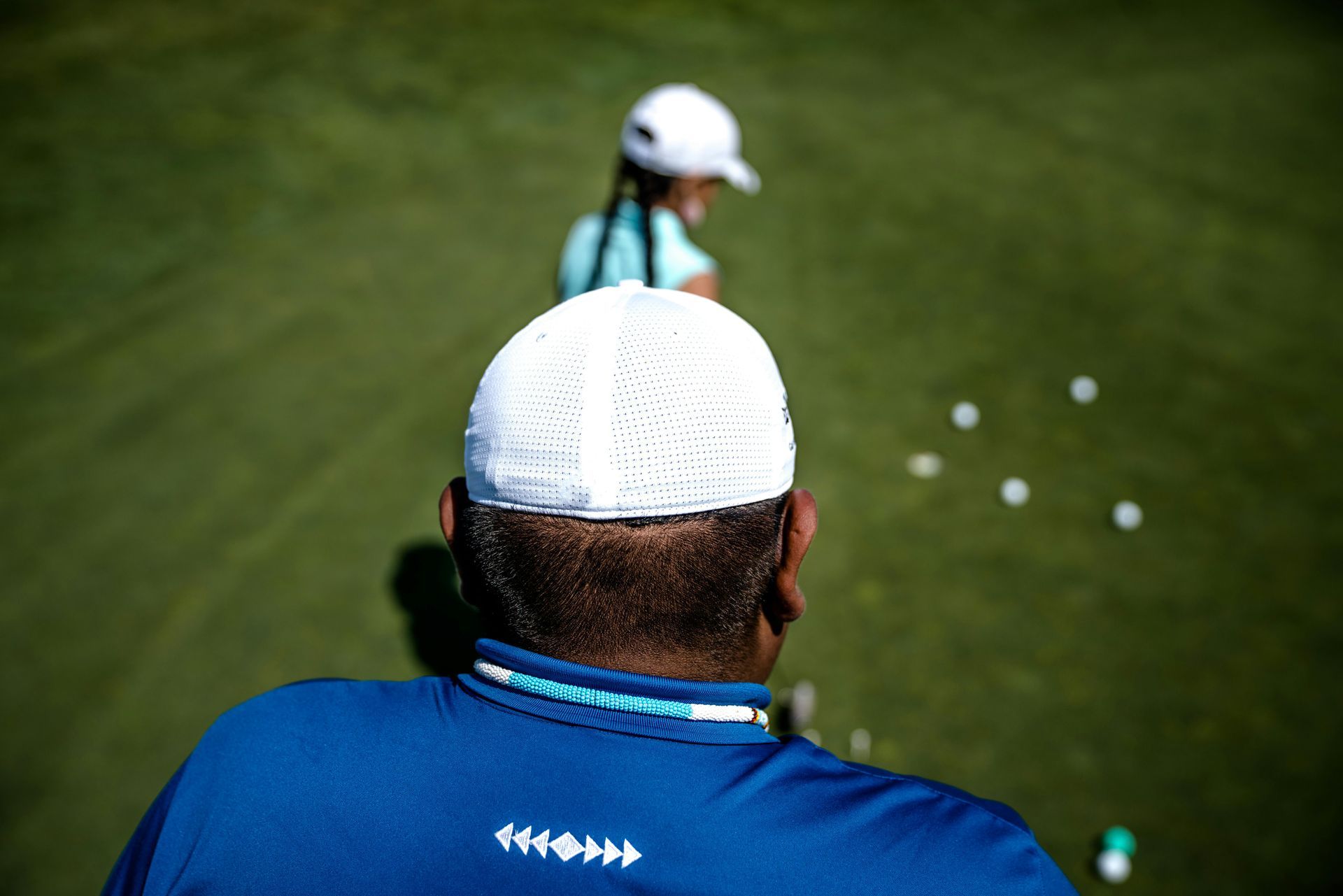 A man in a blue shirt watches a woman in a white hat on a golf green with golf balls.