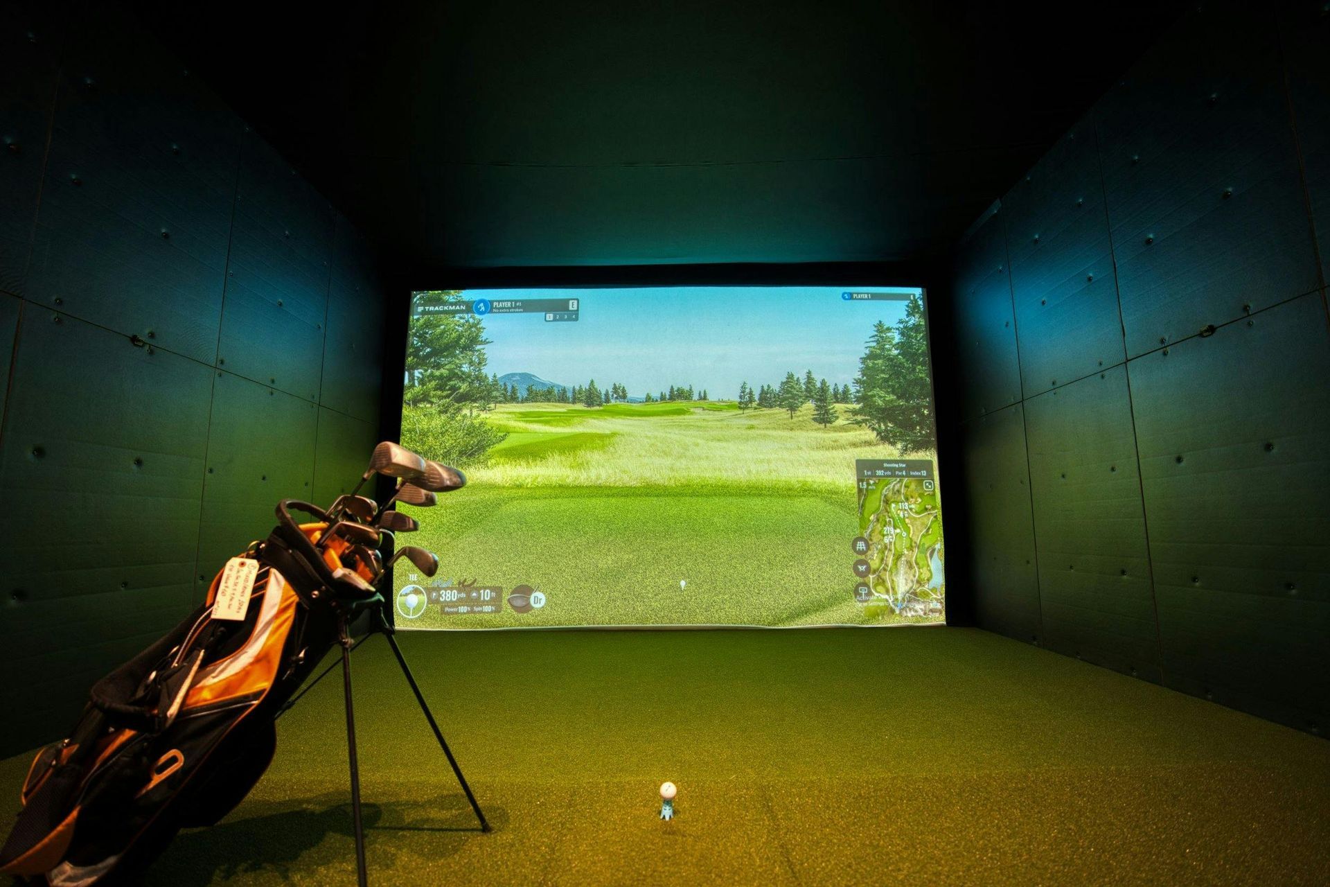 Golf simulator setup: golf bag, clubs, ball on turf, screen displaying a golf course.