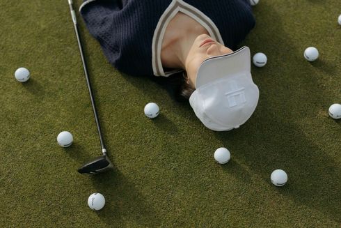 Person on golf course surrounded by golf balls and club, wearing a white hat.