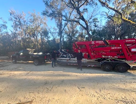 Two people stand near a truck towing a red industrial machine on a trailer in a wooded area.