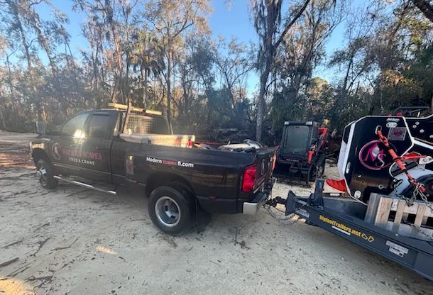 Dark pickup truck towing a trailer with equipment, parked on dirt in a wooded area.