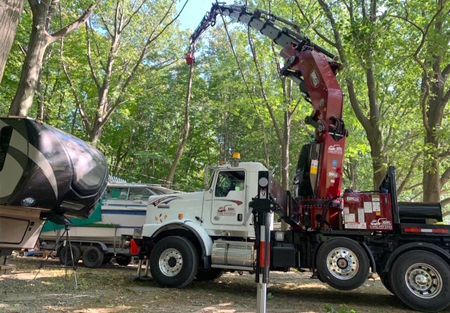 A crane lifting a boat from a trailer, outdoors. Red crane arm, white truck, green trees.