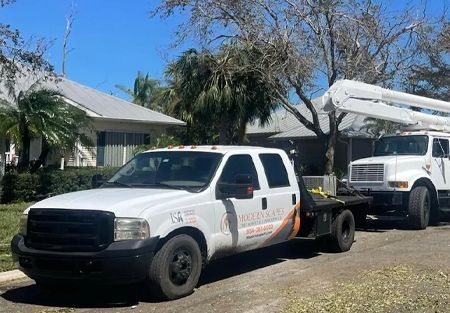 White flatbed truck and boom truck parked in front of a house, possibly for tree trimming.