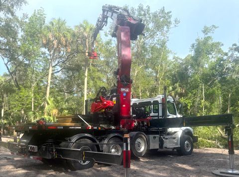 A red crane on a flatbed truck lifting material near trees on a sunny day.