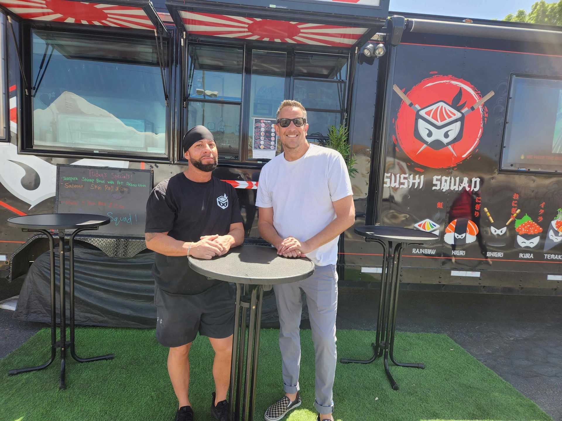 Two men are standing in front of a sushi truck.