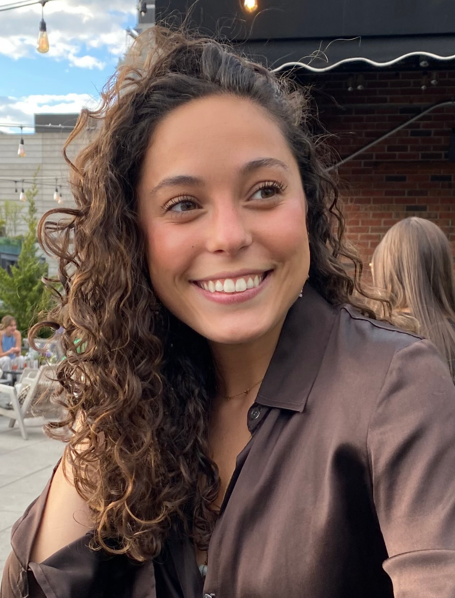 Woman with curly brown hair smiles outdoors in brown shirt.
