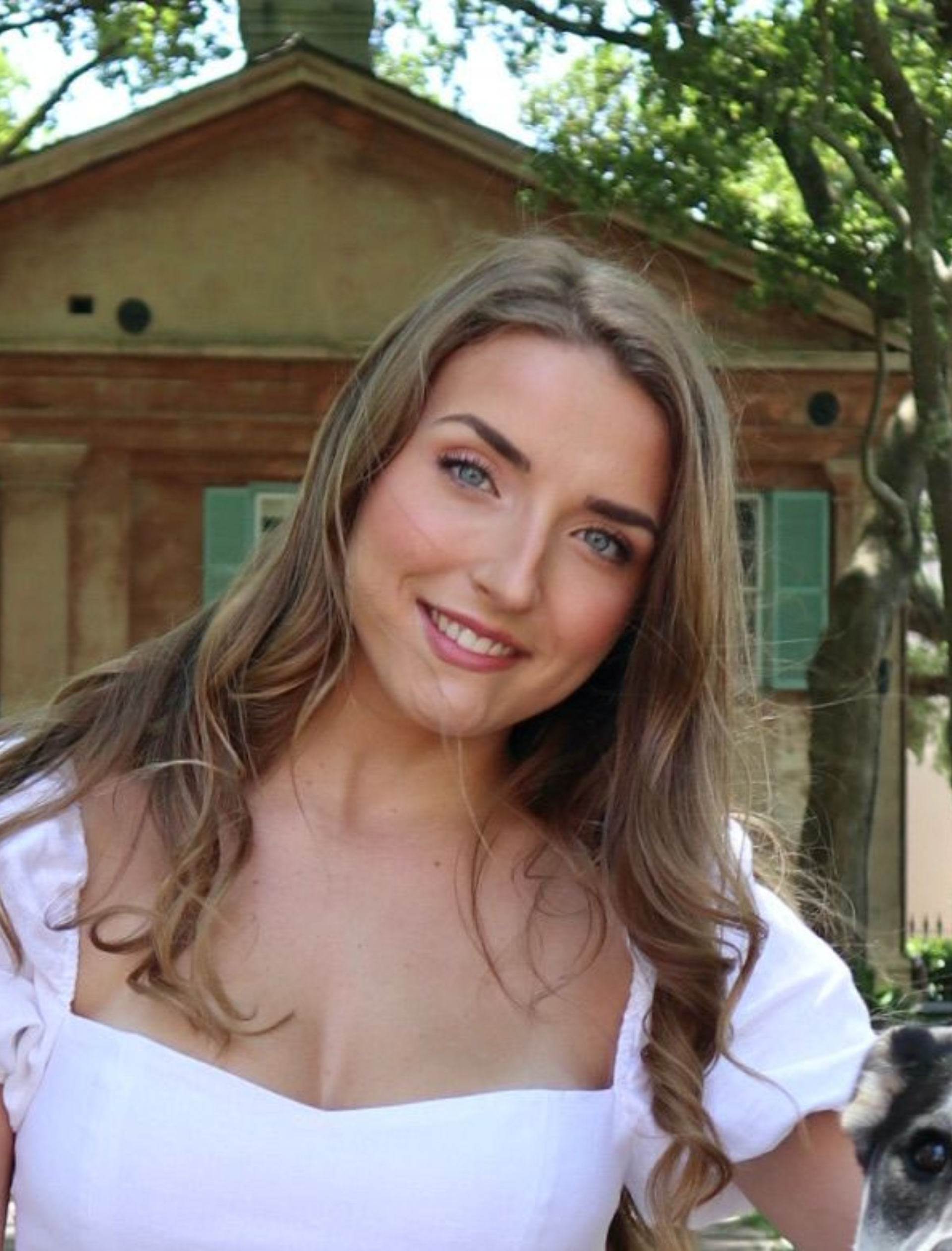 Woman smiles, light brown hair, white top, in front of a building with green shutters.