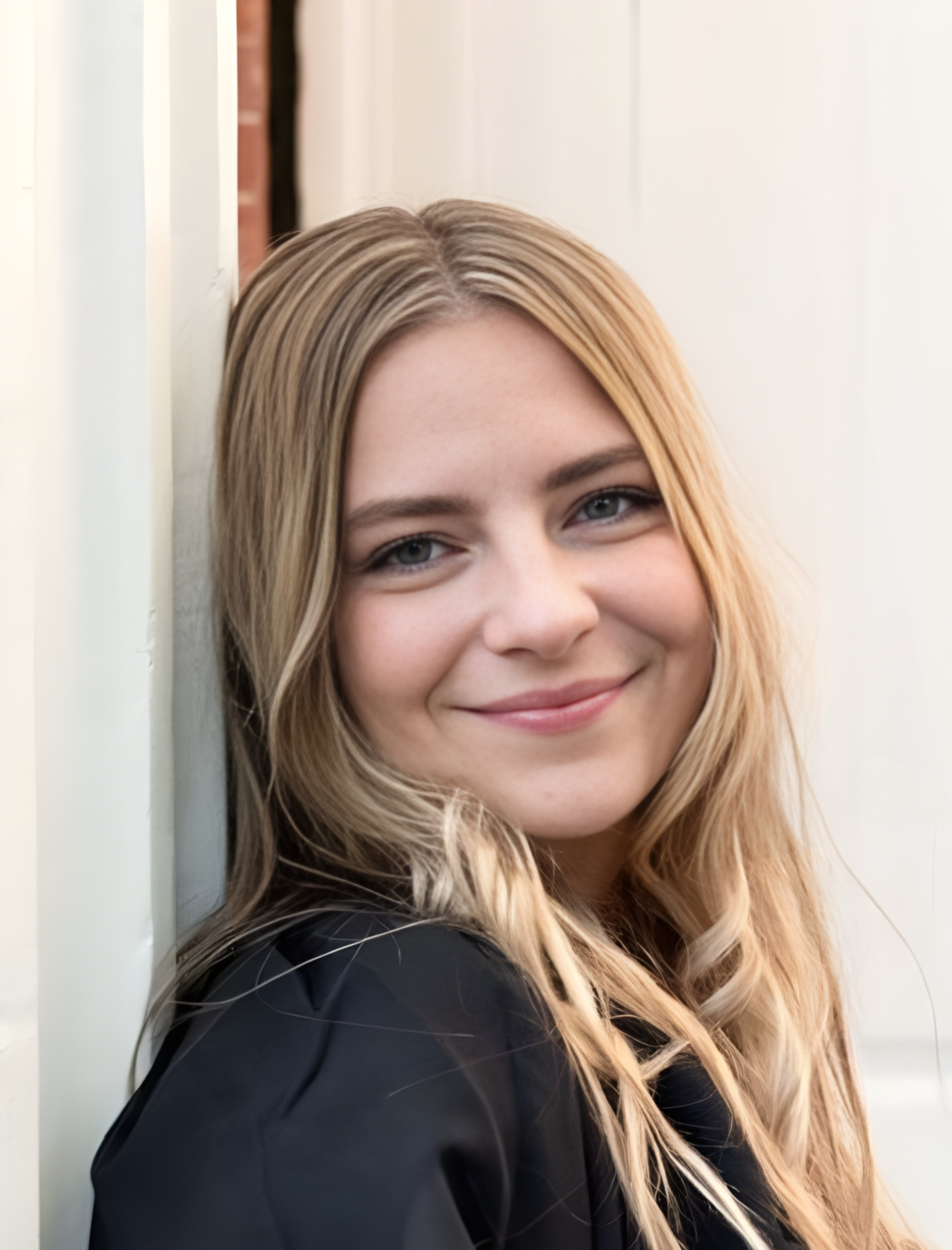 Blond woman smiling, leaning against a white wall outdoors, wearing a black top.