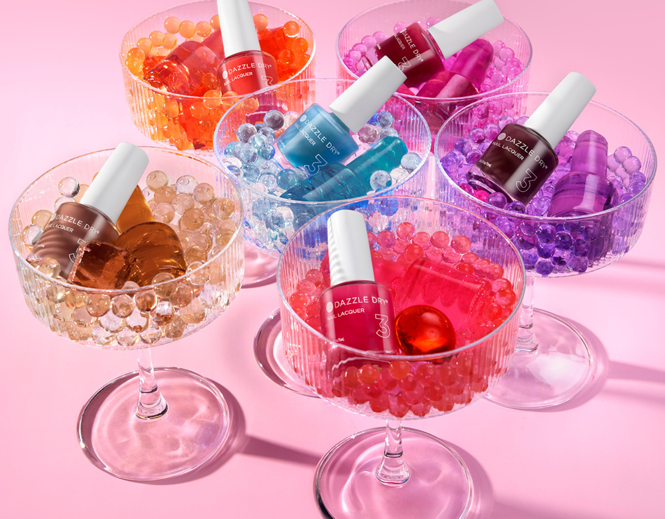 Nail polish bottles in clear bowls filled with colorful gel beads on a pink surface.