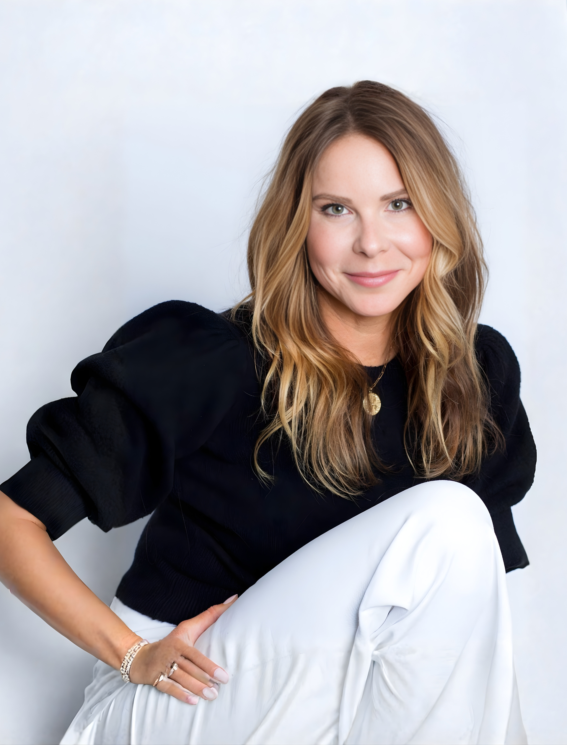 Woman in black sweater and white pants, leaning, smiling.