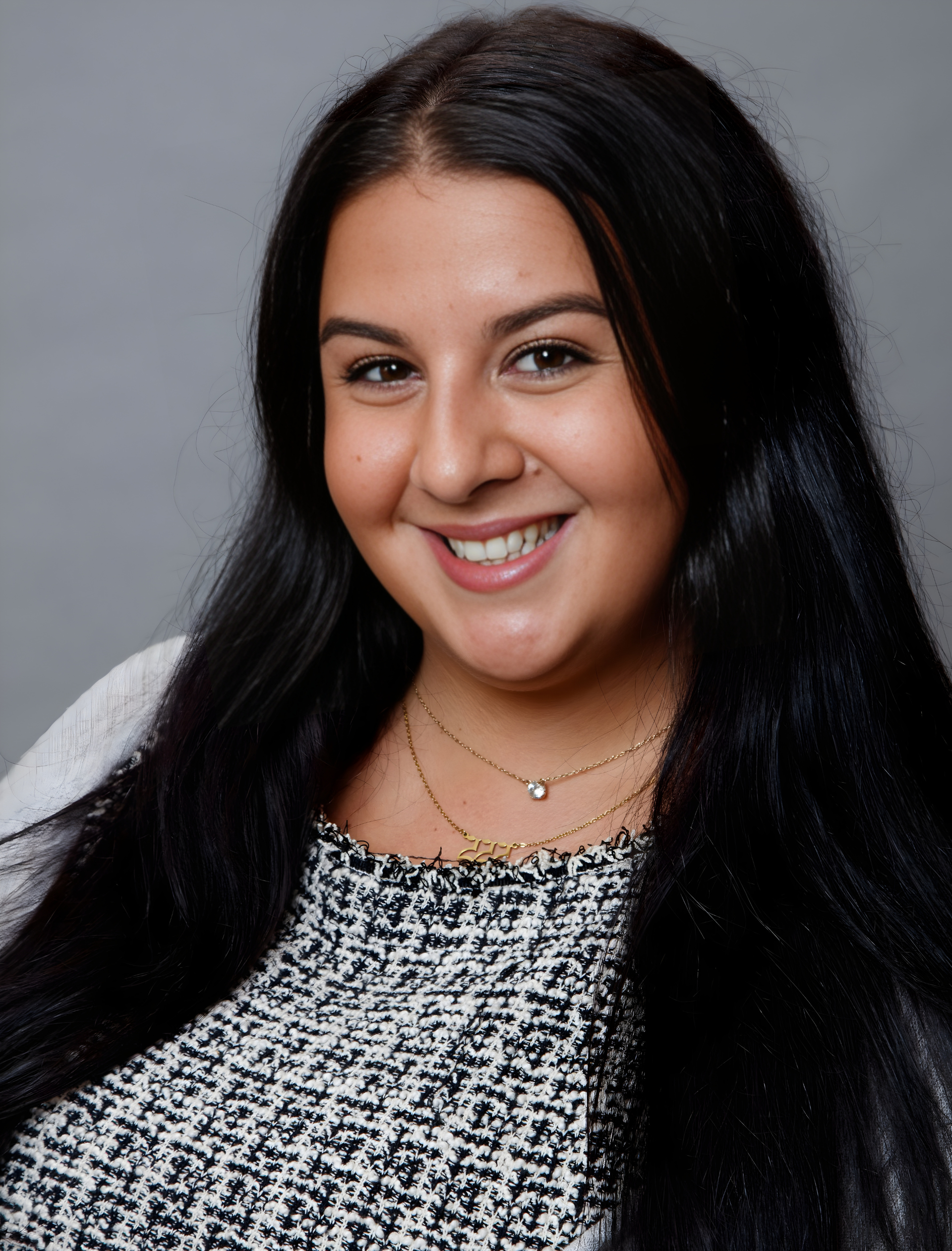 Woman with long, dark hair smiles. She wears a patterned top and gold necklaces. Gray background.