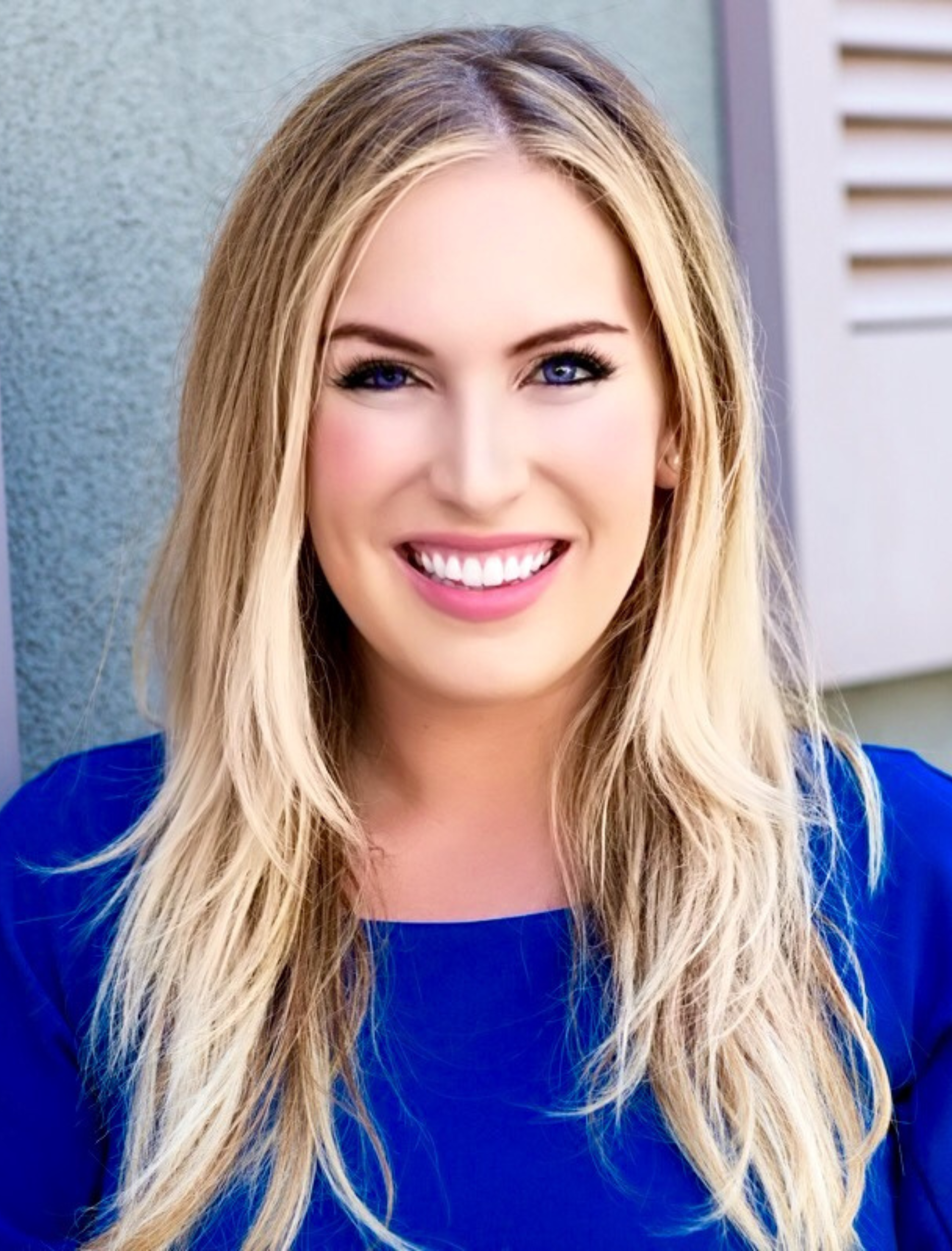 Blonde woman in blue smiling at the camera, sunlight, outdoor setting.