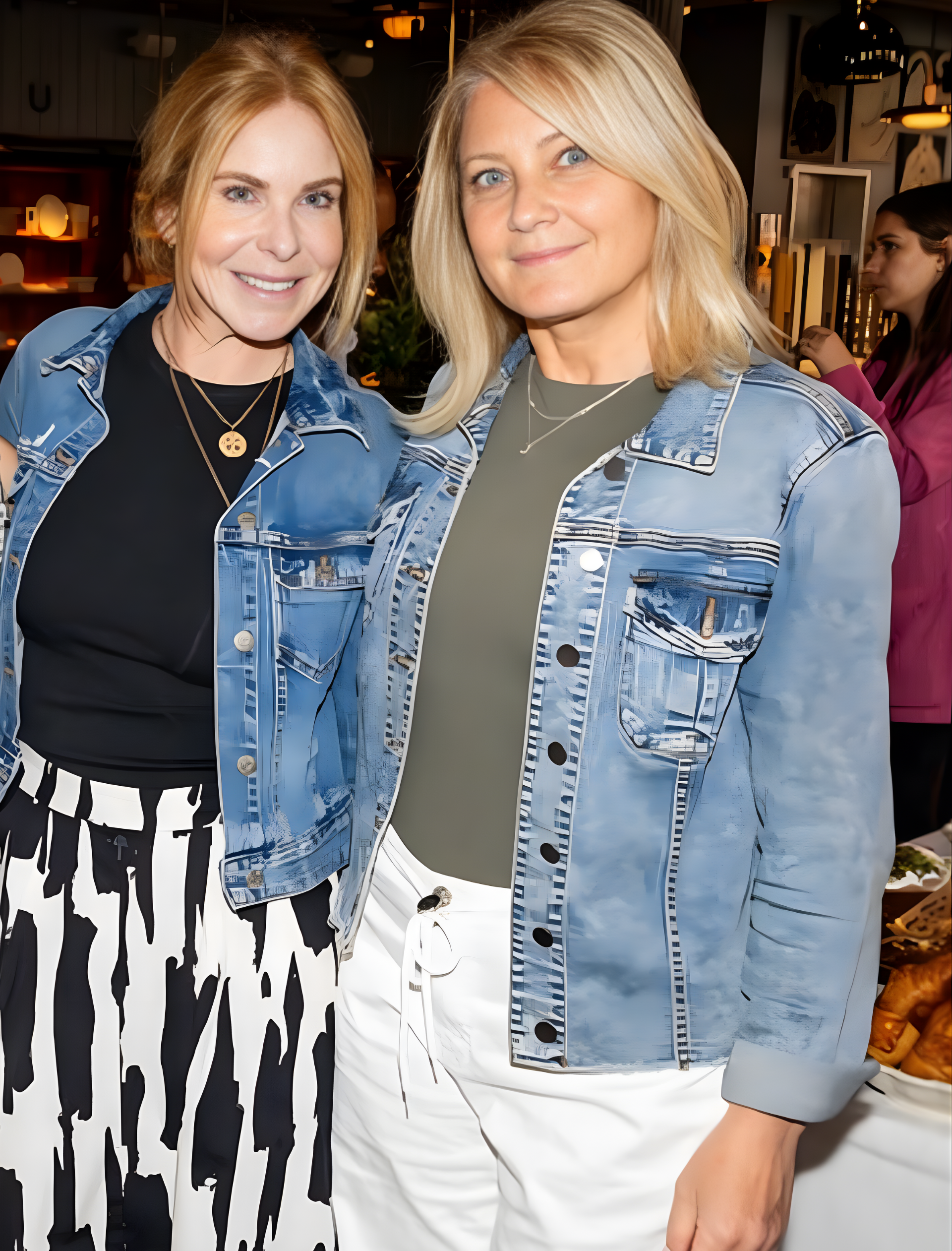 Two women in denim jackets. One wears a black top and patterned skirt; the other, an olive top and white pants.