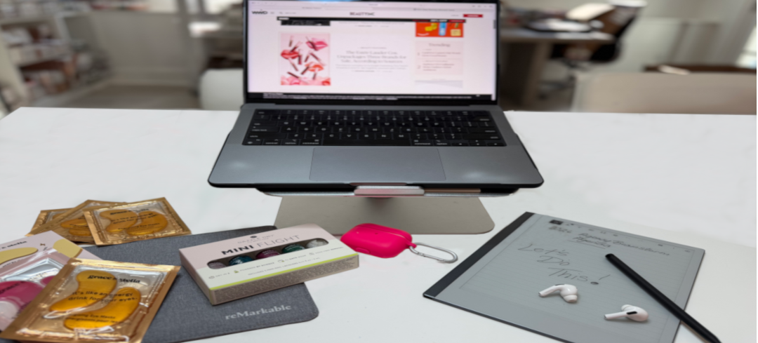 Laptop on stand with skincare products, digital notepad, and headphones on a white desk.