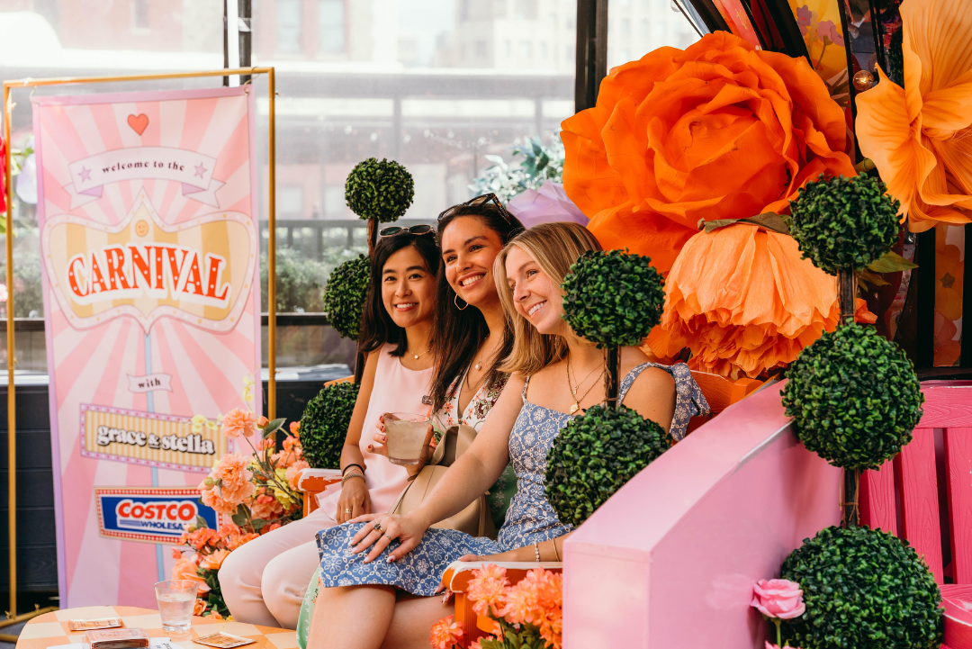 Three smiling women posing in front of a colorful carnival-themed backdrop, surrounded by flowers.