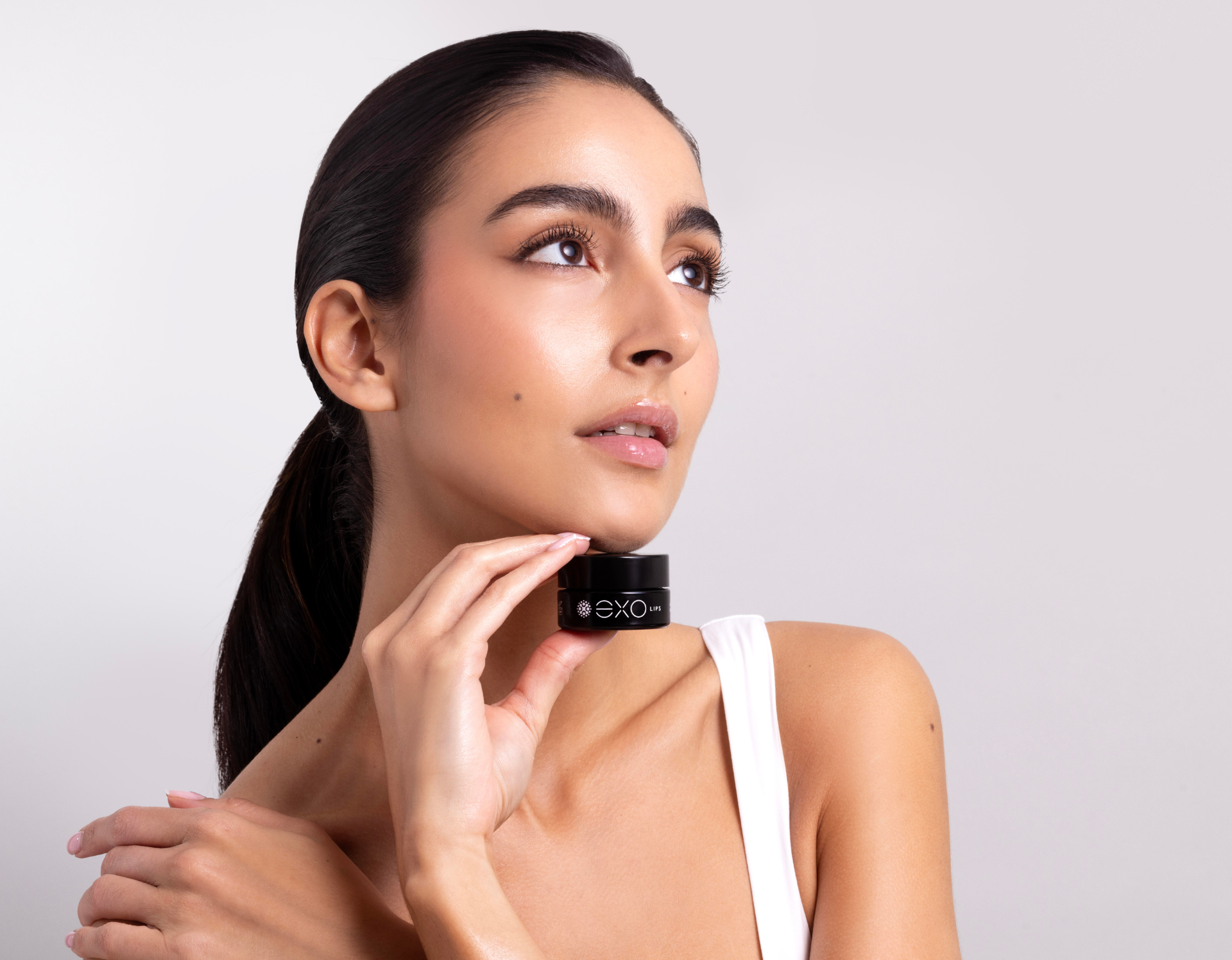 Woman holding a small black jar, looking upward. She wears a white tank top, has dark hair pulled back.